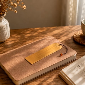 Puede incluir: Un marcapáginas rectangular de metal dorado con acabado cepillado y marcas de regla grabadas en un borde. El marcapáginas presenta un patrón de filigrana decorativo en la parte superior y está unido a un cordón negro con una cuenta de color ámbar.