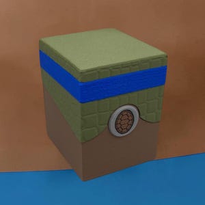 Peut inclure: Une boîte de rangement rectangulaire avec une base marron et une partie supérieure vert olive texturée. Une bande horizontale bleu vif entoure la partie supérieure. Un emblème circulaire avec un motif de carapace de tortue est centré.