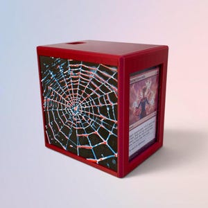 Puede incluir: Una caja de almacenamiento rectangular roja con un panel lateral negro que presenta un diseño de telaraña en relieve en blanco y azul. El panel frontal tiene una ventana rectangular vertical que muestra una carta coleccionable.