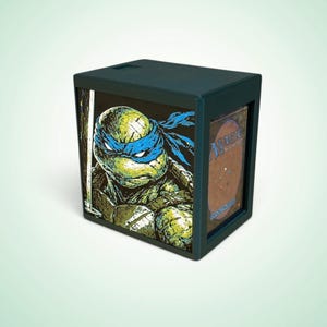 May include: A dark-coloured rectangular card storage box featuring a graphic of a green turtle character wearing a blue mask and holding a sword. The side of the box displays a Magic The Gathering card with a brown back and five coloured mana symbols.