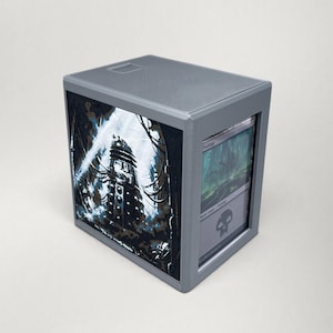 Könnte beinhalten: Eine graue, rechteckige Aufbewahrungsbox mit strukturierter Oberseite. Die Seitenwand zeigt eine schwarz-weiße Illustration einer Roboterfigur mit Kabeln, während die Vorderseite eine Swamp-Sammelkarte mit einem Totenkopfsymbol zeigt.