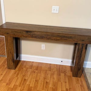 Könnte beinhalten: Ein rechteckiger Holztisch mit dunkelbraunem Finish. Der Tisch hat eine dicke Oberseite und zwei stabile, rechteckige Beine. Die Holzmaserung ist sichtbar und verleiht Textur. Der Tisch ist etwa 152 cm lang.