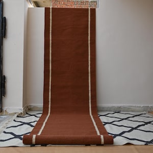 May include: A long, rectangular runner rug in a solid deep rust-brown color featuring two vertical cream-colored stripes running the length of the piece. The runner is draped over a white, cream, and black diamond-patterned area rug with fringe.