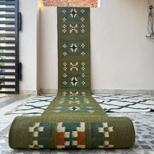 May include: A long, olive green runner rug featuring a repeating geometric pattern in cream, dark blue, and burnt orange. The rug is partially rolled at the front, displaying a thick, woven texture with symmetrical, block-style motifs.