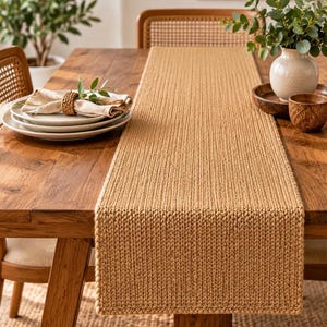 May include: A rectangular table runner featuring a thick, chunky knit texture in a solid tan colour. The runner is laid flat across the centre of a wooden dining table, extending from one end to the other with a consistent, uniform stitch pattern.