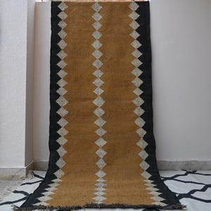 Puede incluir: Una alfombra rectangular larga con un panel central de color amarillo mostaza flanqueado por bordes negros. Una fila vertical de diamantes blancos recorre el centro, mientras que diamantes blancos también bordean los límites entre el amarillo y el negro.