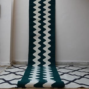 Puede incluir: Una alfombra de pasillo larga y rectangular con un llamativo patrón de zigzag en chevron de color verde oscuro y blanco roto que recorre verticalmente toda su longitud. La alfombra cuelga de una pared y está enrollada en el borde inferior.