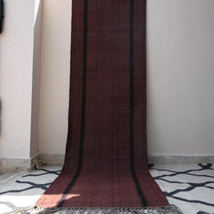 Peut inclure: Un long tapis de couloir rectangulaire d'une couleur marron foncé unie, orné de deux bandes verticales noires sur toute la longueur. Le tapis présente un aspect tissé texturé et est terminé par une courte frange de couleur claire à la base.