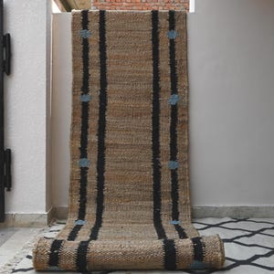Peut inclure: Un long tapis de couloir rectangulaire en fibres naturelles tissées de couleur beige. Le tapis présente deux bandes verticales noires proéminentes sur toute sa longueur, agrémentées de petits motifs circulaires bleu clair le long des lignes.