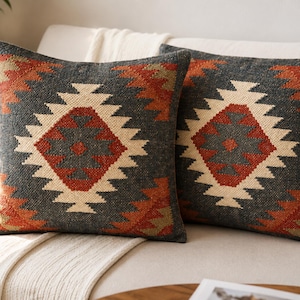 May include: Two square throw pillows featuring a dark charcoal gray base with a prominent geometric diamond pattern in cream, burnt orange, and muted olive green. The woven texture displays a repeating stepped motif across the front surface.