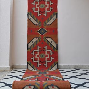 Puede incluir: Una alfombra pasillera larga y rectangular con una base de color rojo vibrante y patrones geométricos en negro, blanco, gris y amarillo mostaza. El diseño incluye motivos repetitivos de diamantes y ángulos, con un extremo enrollado en cilindro.
