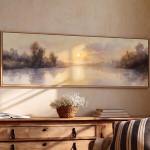 May include: A panoramic landscape painting in a light wood frame depicting a sunset over a calm river with silhouetted trees. The artwork hangs above a long wooden sideboard featuring three drawers with black metal pulls and a white pitcher.