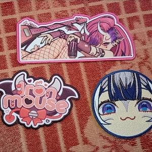 Puede incluir: Tres parches bordados sobre una superficie roja estampada. El parche superior es rectangular con un borde rosa, con un personaje de dibujos animados con pelo y cuernos rosas. El parche del medio dice "Iron Mouse" en rosa y blanco. El parche inferior es redondo con una cara azul y blanca de estilo anime.