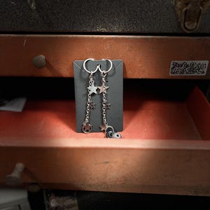 May include: Silver dangle earrings with star charms and a small black and silver accent. The earrings are displayed on a black card against a backdrop of a red and black toolbox.