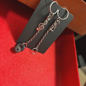 K&ouml;nnte beinhalten: Silberfarbene Ohrringe mit Noten-Anh&auml;ngern und einer daran h&auml;ngenden Kette. Ein Ohrring hat einen Violinschl&uuml;ssel-Anh&auml;nger, der andere einen Noten-Anh&auml;nger. Jeder Ohrring hat einen grauen Ohrh&ouml;rer. Die Ohrringe werden auf einer schwarzen Karte pr&auml;sentiert.