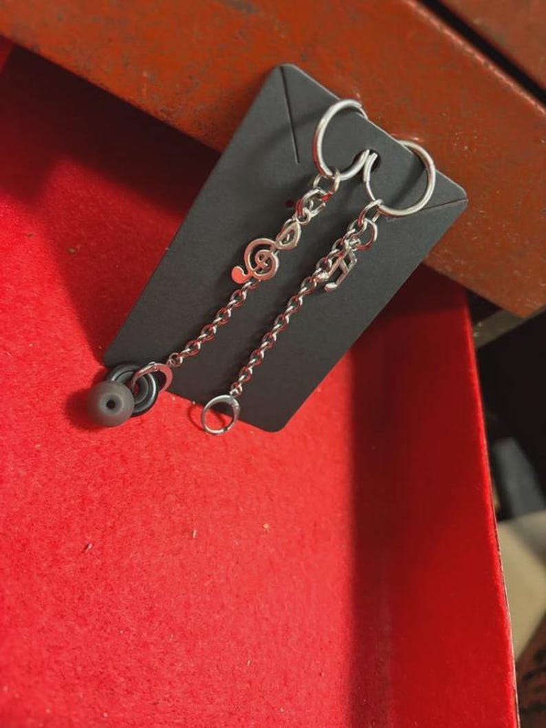K&ouml;nnte beinhalten: Silberfarbene Ohrringe mit Noten-Anh&auml;ngern und einer daran h&auml;ngenden Kette. Ein Ohrring hat einen Violinschl&uuml;ssel-Anh&auml;nger, der andere einen Noten-Anh&auml;nger. Jeder Ohrring hat einen grauen Ohrh&ouml;rer. Die Ohrringe werden auf einer schwarzen Karte pr&auml;sentiert.