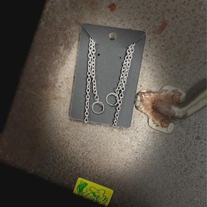 May include: Silver chain earrings with a small hoop and dangling chain design. The earrings are displayed on a dark gray card against a textured, metallic surface. A small, yellow candy wrapper with green graphics is visible.