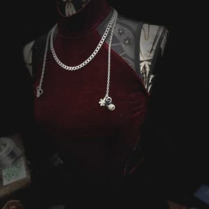 Op de afbeelding: Bordeaux fluwelen halterjurk op een mannequin, gecombineerd met een zilveren ketting. De ketting heeft een dikke schakel en delicate bedels. De mannequin heeft een patroon op de hals en schouders, tegen een donkere achtergrond.