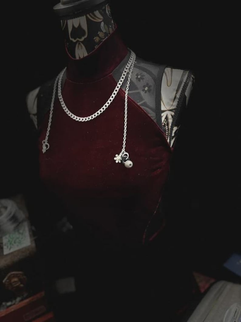 Op de afbeelding: Bordeaux fluwelen halterjurk op een mannequin, gecombineerd met een zilveren ketting. De ketting heeft een dikke schakel en delicate bedels. De mannequin heeft een patroon op de hals en schouders, tegen een donkere achtergrond.