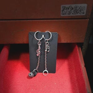K&ouml;nnte beinhalten: Silberne Ohrringe mit Noten-Anh&auml;ngern und h&auml;ngenden Ketten. Ein Ohrring hat einen Violinschl&uuml;ssel, der andere eine Note. Ein Ohrring hat einen schwarzen Ohrh&ouml;rer, der andere einen kleinen Silberring.
