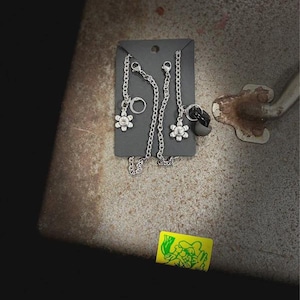 Puede incluir: Un par de cadenas de metal plateado sujetas a una tarjeta de presentación negra. Cada cadena presenta un pequeño dije en forma de flor con un diseño de cara sonriente en el centro. Una cadena incluye un anillo y la otra un auricular gris.