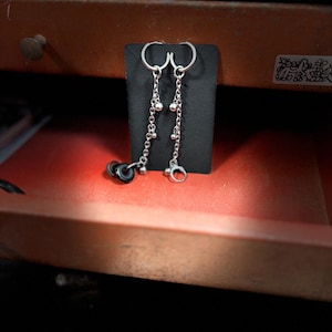 May include: Silver earrings with a chain design. Each earring has a small hoop, a chain of small beads, and a larger charm. The earrings are displayed on a black stand. The earrings are presented on a black stand.