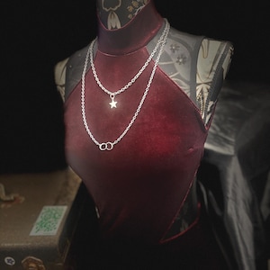 May include: A dark red velvet high-neck top with black mesh side panels, worn with a layered silver-tone chain necklace featuring a small star charm and two interlocking circular rings, displayed on a mannequin against a dark background.