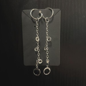 May include: Silver-toned dangle earrings with a chain design. Each earring features a circular hoop at the top, a chain with small charms, and a larger circular charm at the bottom. The earrings are displayed on a black card.