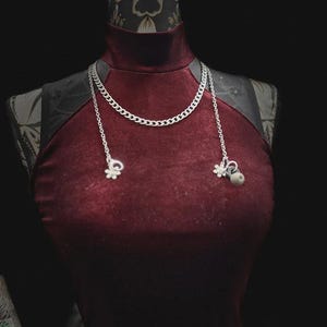 Op de afbeelding: Bordeaux fluwelen jurk met een hoge halslijn en zilveren ketting. De ketting heeft een chunky kettingontwerp met twee bungelende bedels. De jurk wordt gepresenteerd op een mannequin met een zwarte achtergrond.