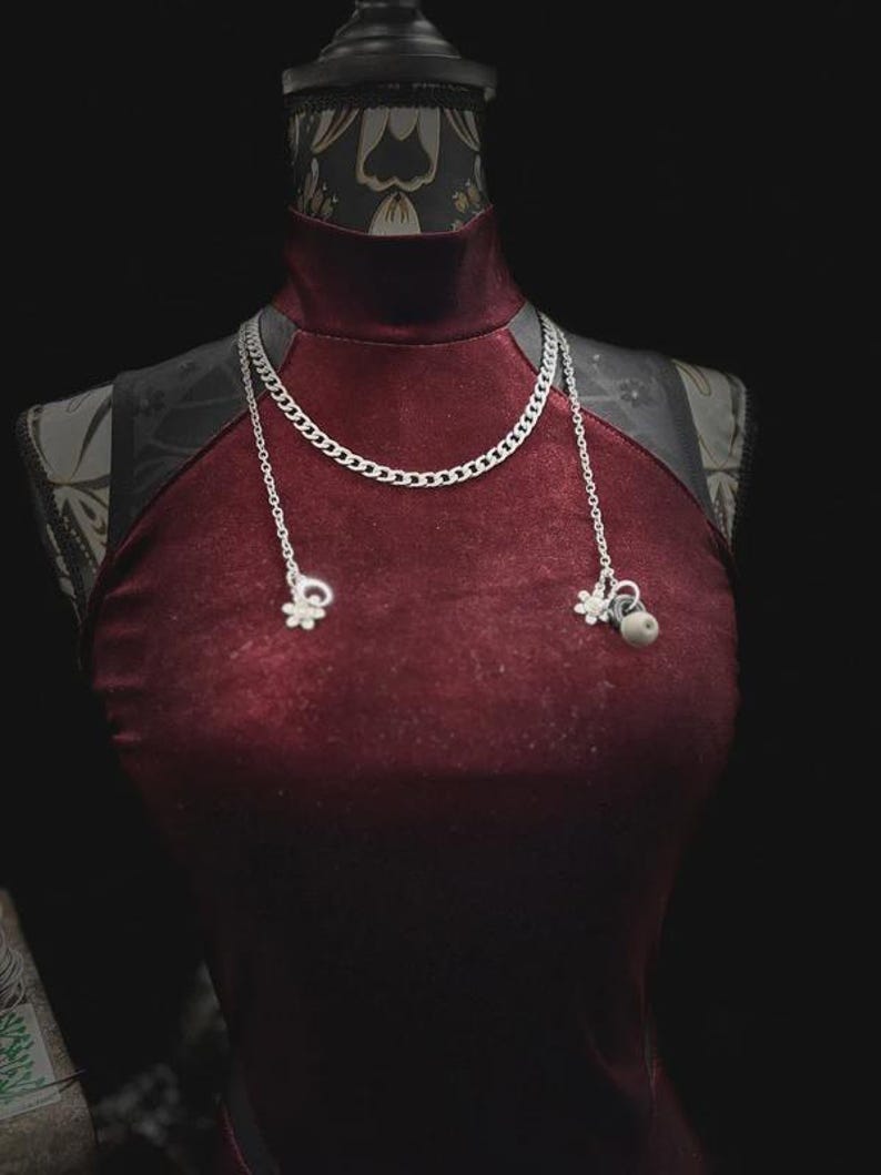 Op de afbeelding: Bordeaux fluwelen jurk met een hoge halslijn en zilveren ketting. De ketting heeft een chunky kettingontwerp met twee bungelende bedels. De jurk wordt gepresenteerd op een mannequin met een zwarte achtergrond.