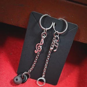 K&ouml;nnte beinhalten: Silberfarbene Ohrringe mit Noten-Anh&auml;ngern. Ein Ohrring zeigt einen Violinschl&uuml;ssel, der andere eine Musiknote. Jeder Anh&auml;nger h&auml;ngt an einer Kette und einem Ring. Die Ohrringe werden auf einer schwarzen Karte vor rotem Hintergrund pr&auml;sentiert.