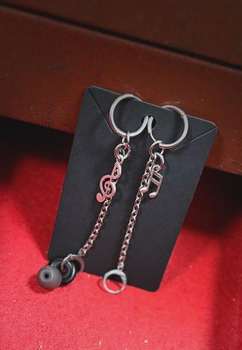 K&ouml;nnte beinhalten: Silberfarbene Ohrringe mit Noten-Anh&auml;ngern. Ein Ohrring zeigt einen Violinschl&uuml;ssel, der andere eine Musiknote. Jeder Anh&auml;nger h&auml;ngt an einer Kette und einem Ring. Die Ohrringe werden auf einer schwarzen Karte vor rotem Hintergrund pr&auml;sentiert.