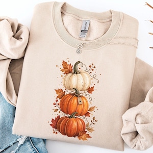 May include: A sand-colored crewneck sweatshirt featuring a graphic print of three stacked pumpkins in shades of white and orange, surrounded by autumn leaves and berries. The sweatshirt has long sleeves and a ribbed collar, with a Gildan brand label.
