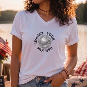 May include: A white V-neck t-shirt featuring a graphic of a disco ball in the center, surrounded by the circular text "RESPECT YOUR MOTHER" in a bold, dark font. The shirt has short sleeves and a standard fit, made of a plain white fabric.
