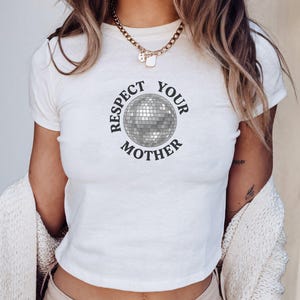 May include: A white short-sleeved t-shirt featuring a graphic of a silver disco ball in the center. The text "RESPECT YOUR MOTHER" is printed in a dark, curved font around the disco ball. The shirt has a crew neckline and a fitted silhouette.