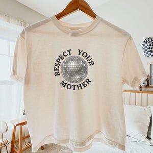 May include: An ivory short-sleeve t-shirt featuring a graphic of a silver disco ball in the center. The text "RESPECT YOUR MOTHER" is printed in black capital letters in a circular arrangement around the disco ball graphic on the front.
