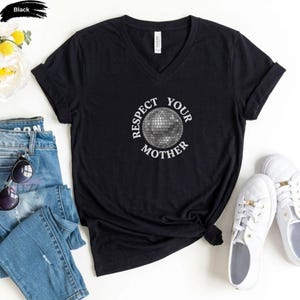 May include: A black V-neck t-shirt featuring a graphic of a silver disco ball in the center, surrounded by the white text "RESPECT YOUR MOTHER" arranged in a circular pattern. The shirt has short sleeves and a standard fit.