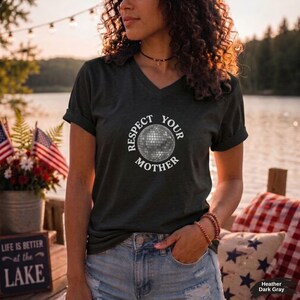 May include: A dark gray V-neck t-shirt featuring a white graphic of a disco ball in the center, surrounded by the circular text "RESPECT YOUR MOTHER" in a bold, white, sans-serif font. The shirt has short sleeves and a standard fit.