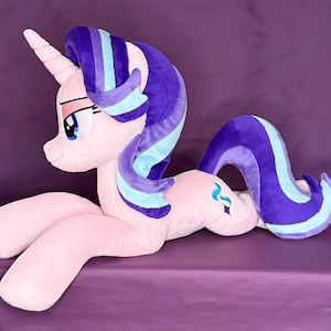 May include: A plush unicorn toy with light pink fur, a spiraled horn, and a long mane and tail featuring deep purple and light blue stripes. The character has large blue eyes with long eyelashes and a small blue and purple cutie mark on its flank.