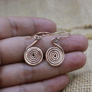 May include: A pair of copper-coloured metal earrings featuring a circular spiral design. Each earring is suspended from a simple curved wire hook, showcasing a polished metallic finish with concentric rings forming the spiral pattern.