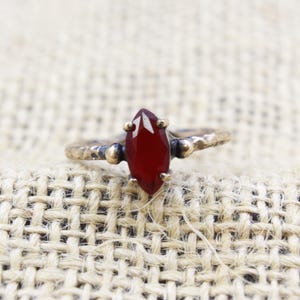May include: A marquise-cut, deep red gemstone ring with a textured, antique gold band. The gemstone is held in place by four prongs. The ring is set against a neutral, woven background.