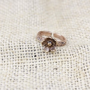 May include: A copper-toned metal ring featuring a five-petaled flower design at the center. The band of the ring has an open, adjustable construction and a textured, rustic finish that extends along the sides of the circular metal band.