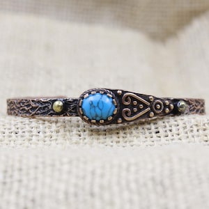 May include: A copper-toned cuff bracelet featuring a textured, branch-like surface. The center displays an oval, bright blue stone with dark veining, flanked by small metallic spheres and decorative scrollwork patterns along the band.