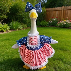 May include: A goose wearing a patriotic outfit. The outfit includes a blue bow with white stars, a dress with a red and white striped skirt, and a blue bodice with white stars. The goose is standing on green grass.