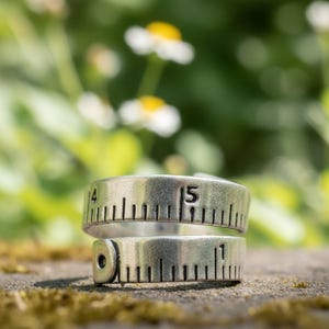 May include: A silver ring designed to look like a measuring tape. The ring has engraved markings and the number 15. The ring is a unique piece of jewellery with a vintage aesthetic.