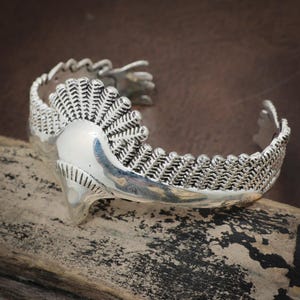 Puede incluir: Un brazalete de plata con un diseño intrincado. El brazalete presenta un elemento central redondeado con una superficie texturizada, flanqueado por patrones detallados en forma de pluma. El diseño abierto facilita su uso.