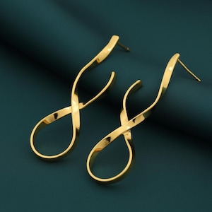 Puede incluir: Un par de pendientes dorados con un diseño abstracto y fluido. Los pendientes tienen una forma retorcida, similar a una cinta, con un poste para orejas perforadas. Los pendientes se muestran sobre un fondo verde azulado oscuro.