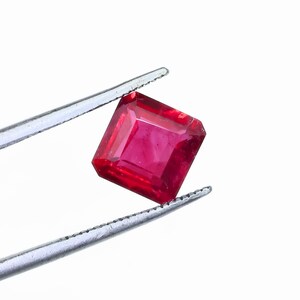 May include: A vibrant, square-cut ruby gemstone held by silver tweezers. The ruby exhibits a deep red hue and a polished surface, reflecting light. The gemstone is set against a plain white background, highlighting its color and clarity.