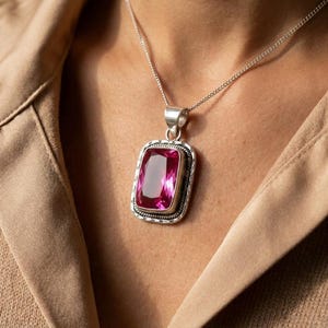 May include: A silver necklace with a rectangular pendant featuring a large, vibrant pink gemstone. The pendant has a decorative silver border and is suspended from a delicate silver chain. The gemstone is a deep, rich pink color.