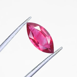 May include: A marquise-cut, faceted ruby gemstone held by tweezers against a white background. The ruby is a vibrant shade of red with visible internal features and sharp, defined facets. The tweezers are silver.